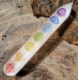 A polished white wand with engraved multicolored chakra symbols, placed on a textured stone surface.