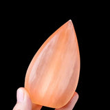 A hand holding a polished orange crystal stone against a black background,. Has a smooth teardrop-shaped crystal.